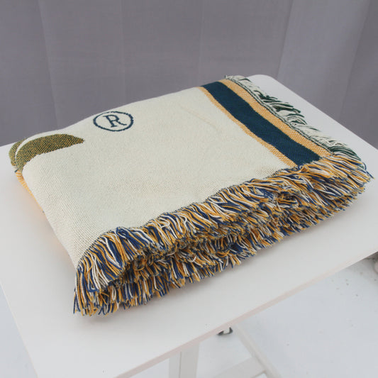 A folded cream-colored woven throw blanket with fringe, featuring a navy blue stripe, a small olive-green leaf motif, and a navy circular logo with an 'R' on the front, displayed on a white table.