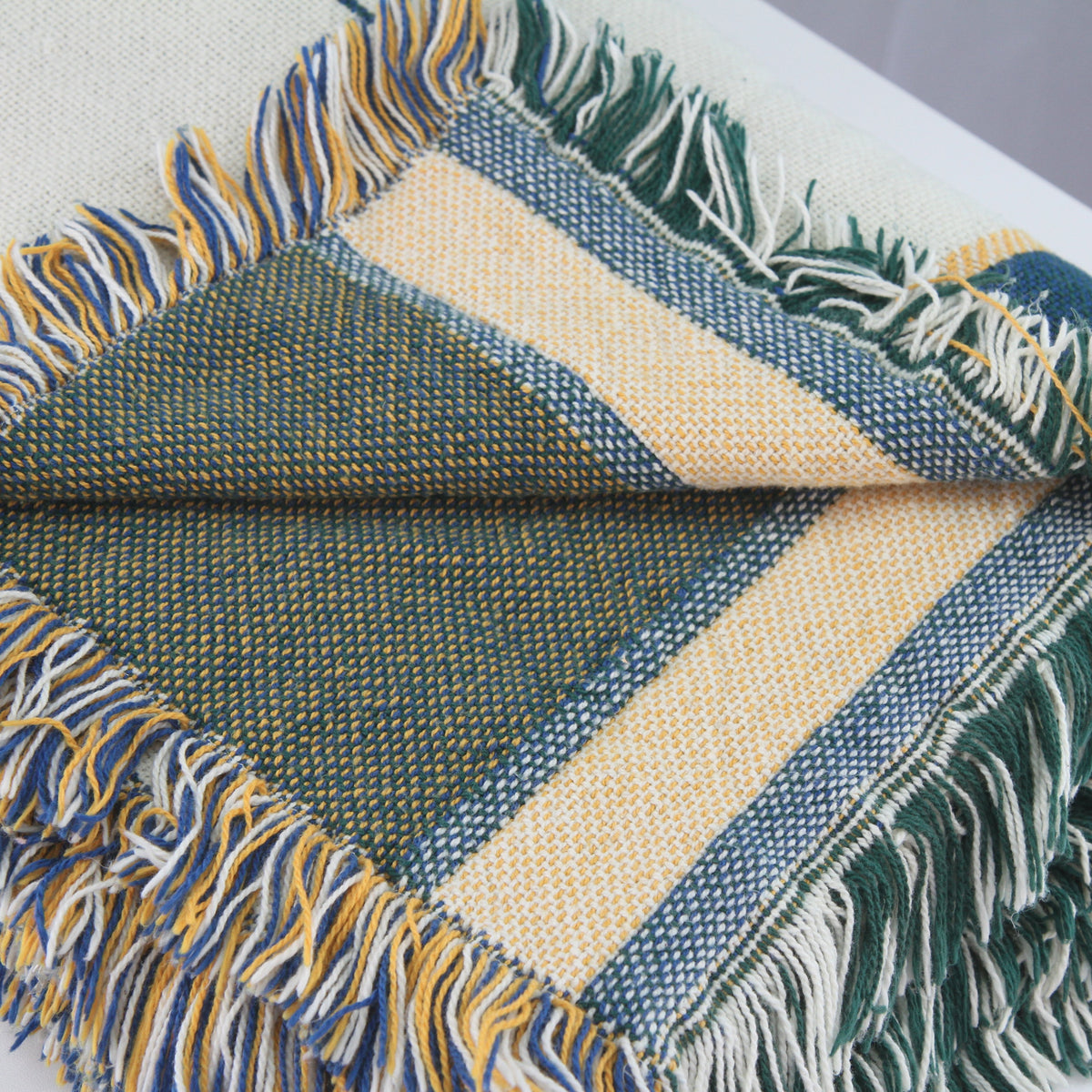 A close-up of a woven fringed scarf or throw blanket with a geometric pattern in dark green, cream, and blue, folded to show its texture and fringe details.