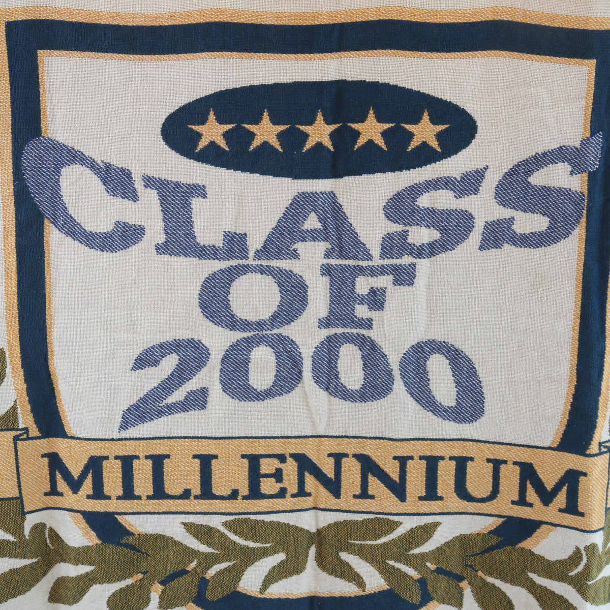 A close-up of a woven textile or tapestry featuring the text 'CLASS OF 2000' and 'MILLENNIUM' in blue lettering on an off-white background, with a navy border, gold stars, and olive-green decorative motifs at the bottom.