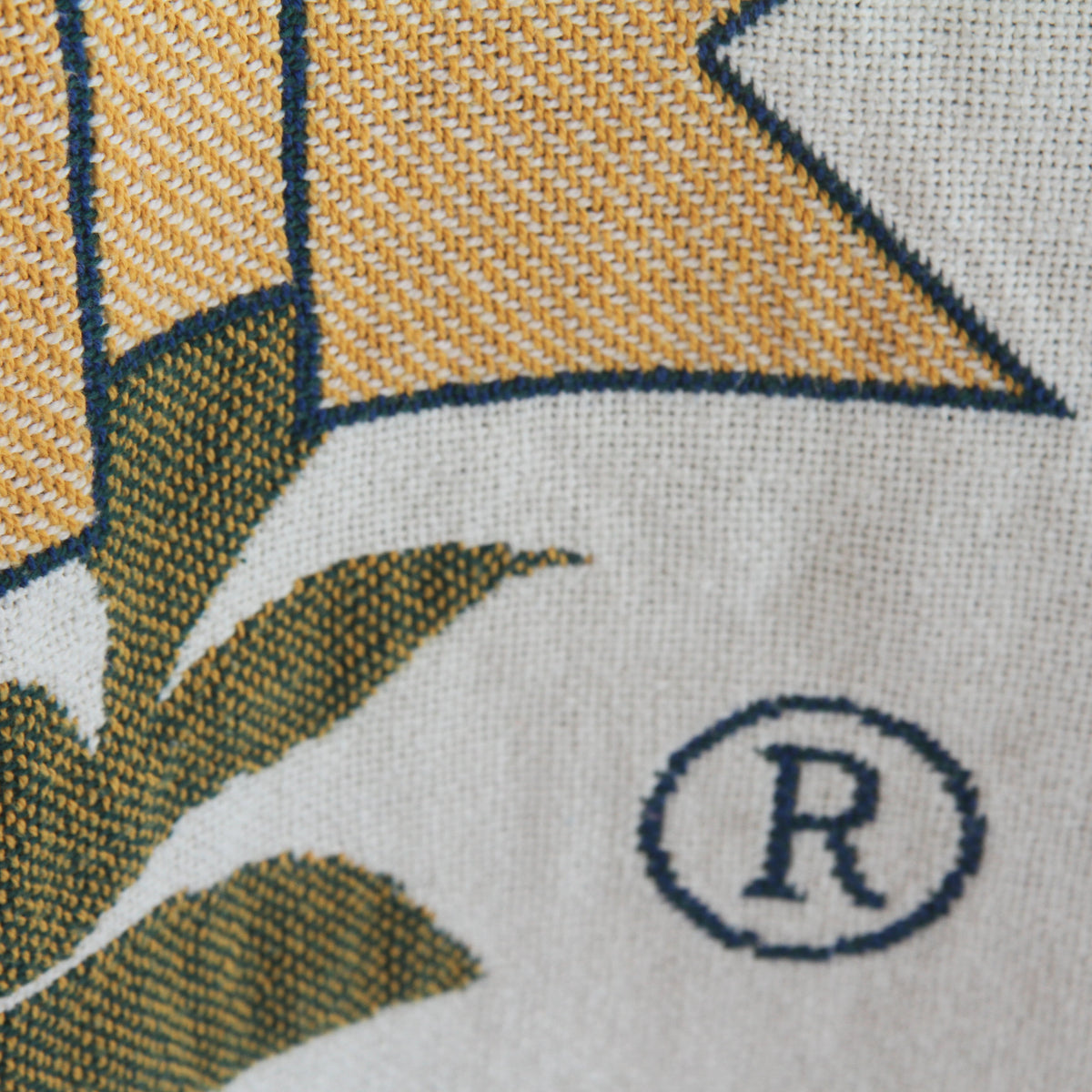 Close-up of a woven textile pattern showing mustard yellow and olive green leaf shapes on an off-white background, with a dark blue registered trademark symbol.