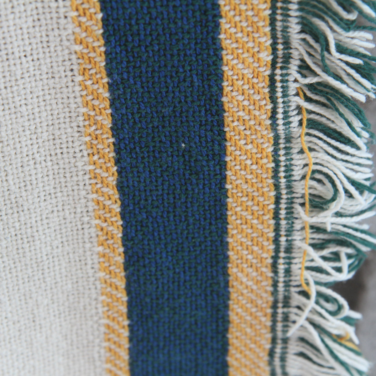 Close-up of a woven textile with vertical stripes in cream, dark blue, mustard yellow, and green, finished with white and green fringe.